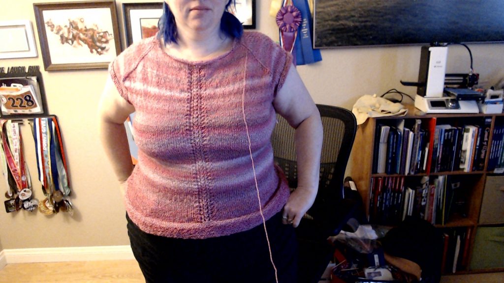 Front view of the pink variegated top, worn by Stephanie, noting the detail of the bottom edge rolling upwards. A single strand of yarn leads off from the neckline, indicating this piece isn't done being knitted.