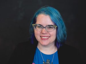 White woman with blue hair and blue catseye glasses, smiling. She wears a blue t-shirt under a black blazer and an octopus necklace.
