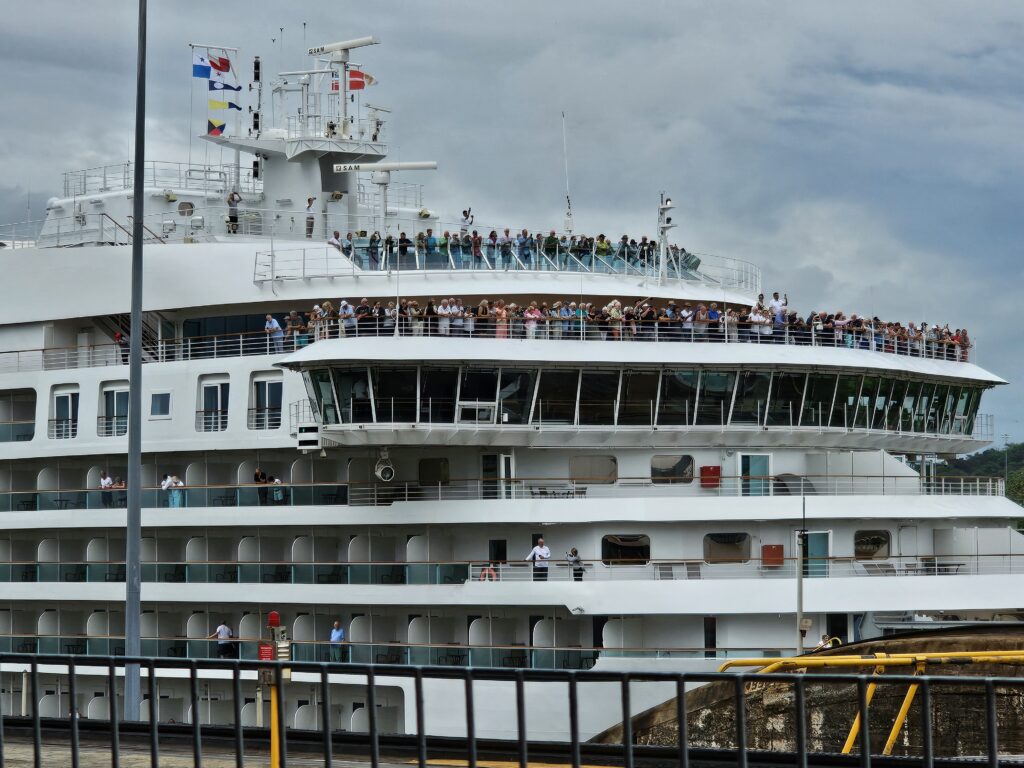 A multi-story cruise ship, with many passengers standing on deck to watch and cheer as the ship is taken through the Panama Canal locks.
