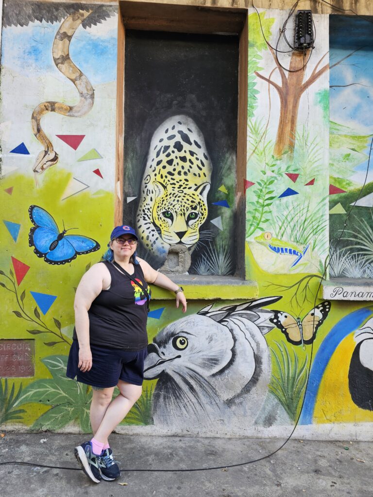 Stephanie, with blue hair, a Cubs hat, and a D&D Pride tank top, standing in front of a colorful mural. The mural has several native animals depicted, including a yellow jaguar, a brown snake, a blue morphos butterfly, an orange monarch butterfly, and a grey bird.