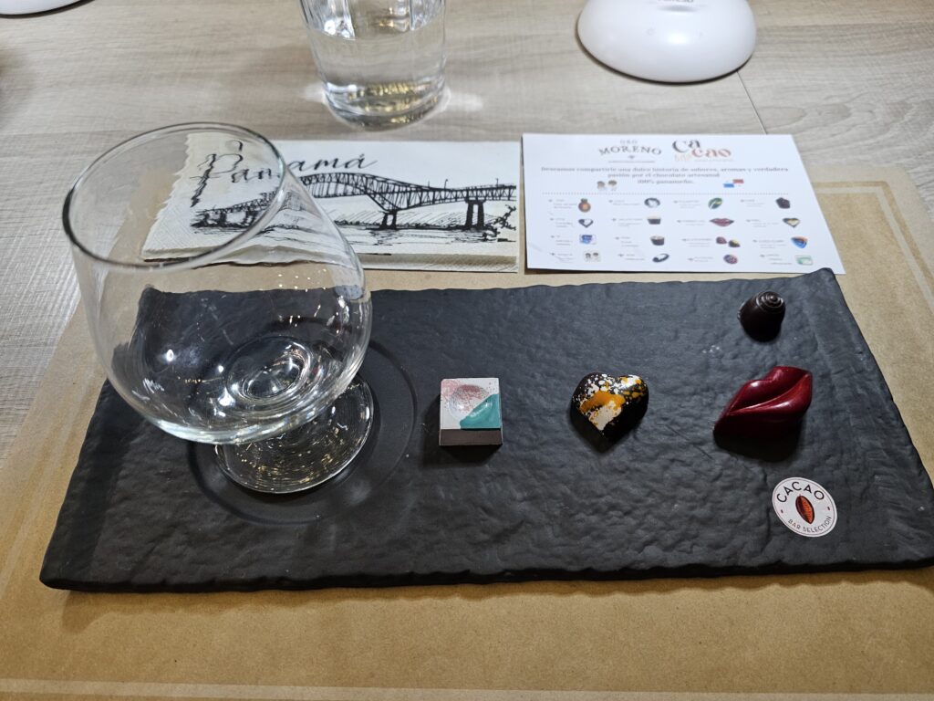 An empty glass and four high quality chocolates are presented on a black plate with a "Cacao" label. Brochures above the plate show the types of chocolate, and a drawing of a bridge with the word "Panama" on it.