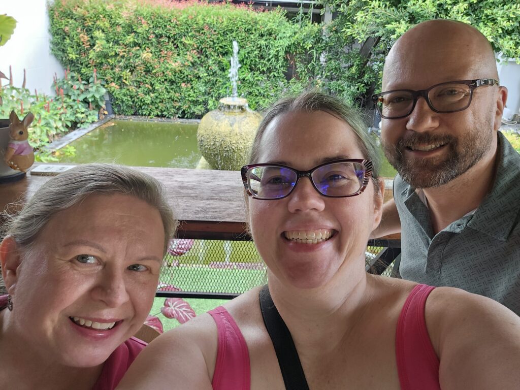 3 smiling white people of middle age-- a blonde woman, a woman with glasses and green hair, and a bearded man in glasses. They are all in front of a garden fountain at a cafe patio.