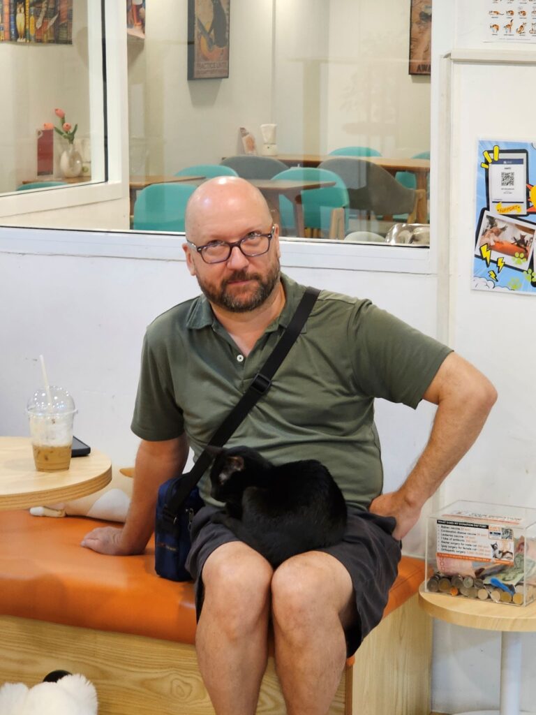 John, a middle aged man with glasses and a beard, seated looking mock-perturbed, at a black kitty that has curled up in his lap. This cat is normally shy with people, but decided John's lap was the best place to be. Introverts recognize each other.