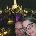 John and Stephanie, a middle-aged male-female white couple, leaning their heads together in a selfie on a ship as it passes the famous Wat Arun, a beautiful temple that is completely lit up with yellow-gold lights at night.