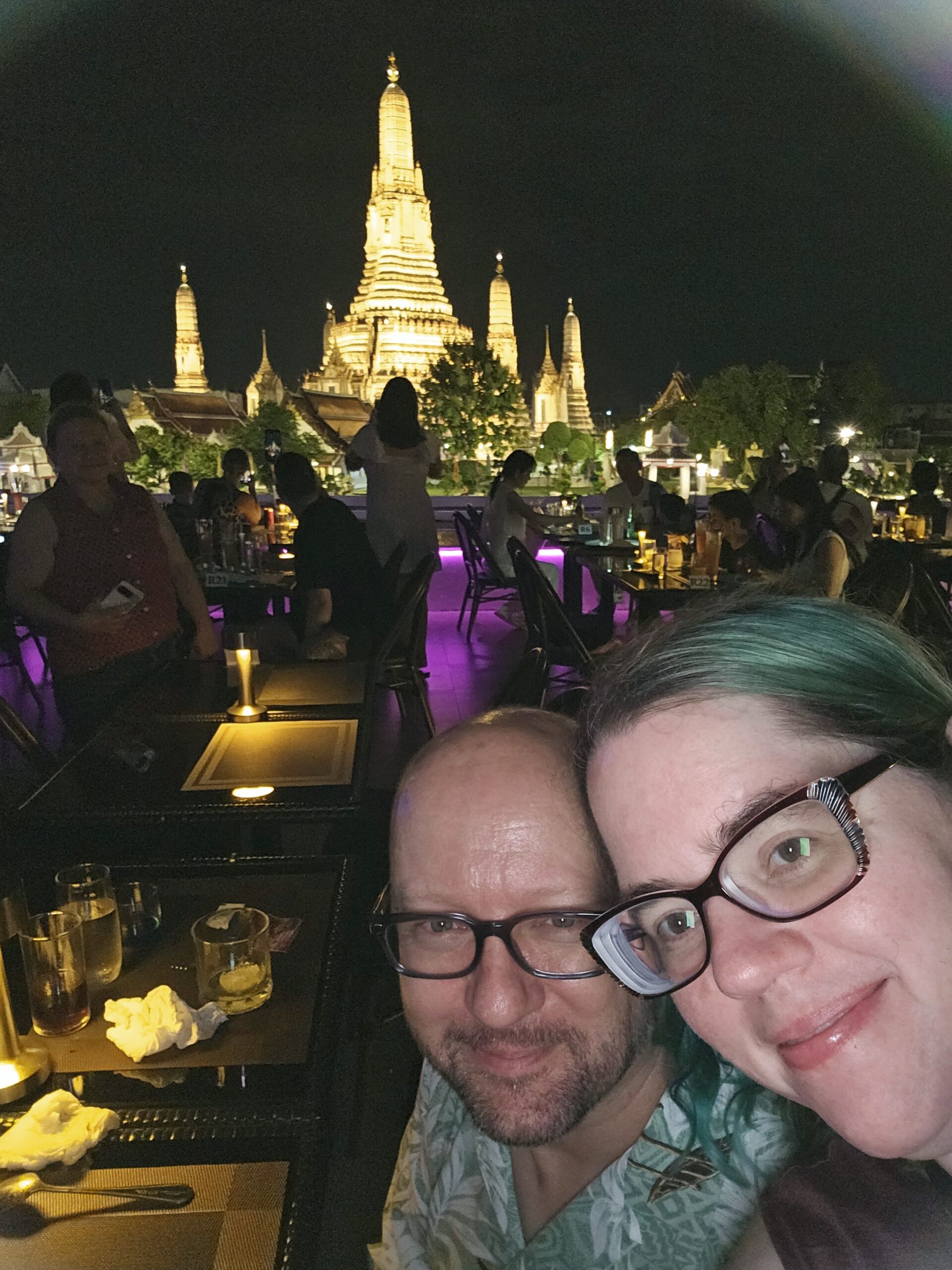 John and Stephanie, a middle-aged male-female white couple, leaning their heads together in a selfie on a ship as it passes the famous Wat Arun, a beautiful temple that is completely lit up with yellow-gold lights at night.