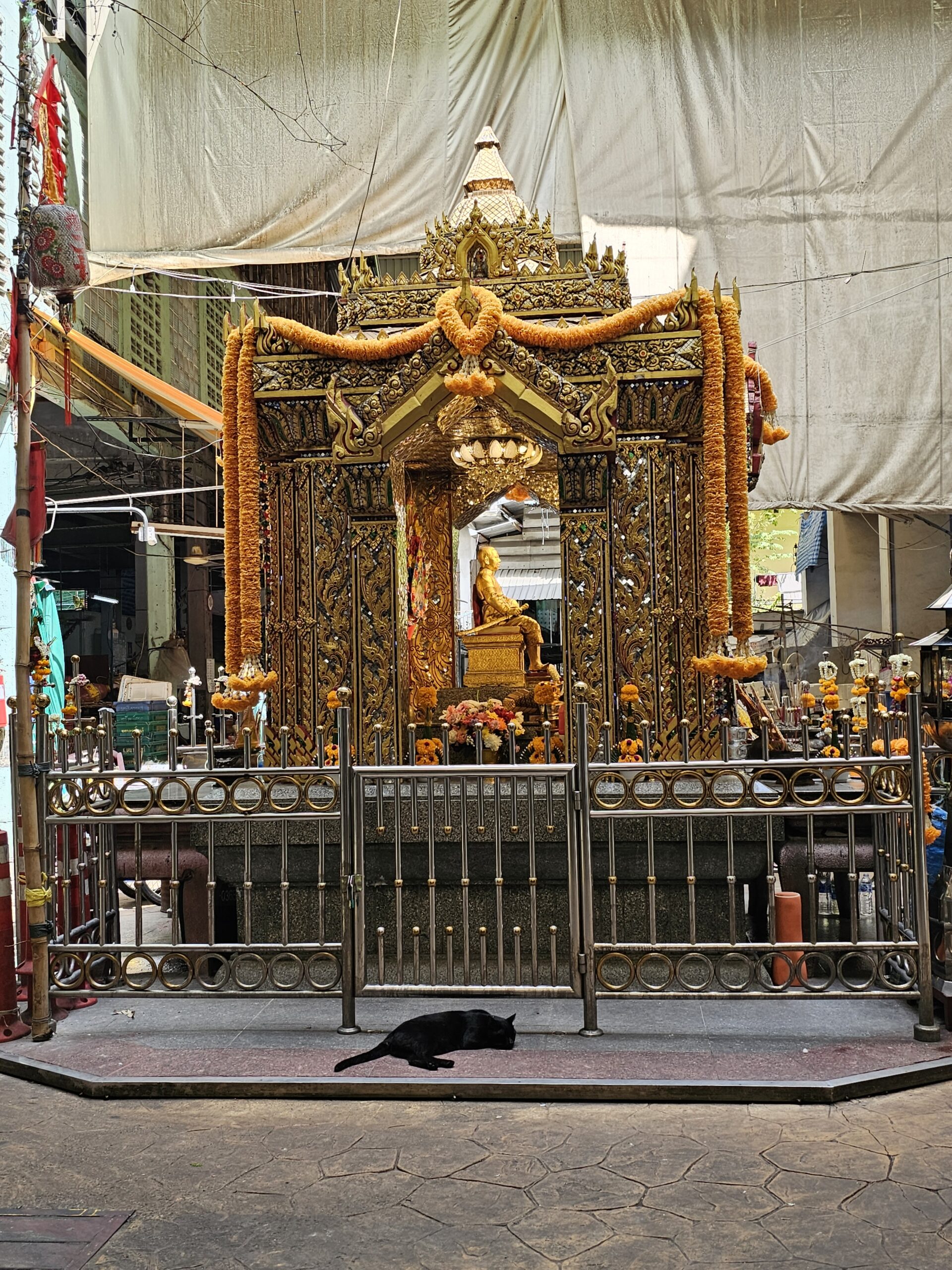 Orange and gold shrine in the center of the flower market. Golden marigold flower garlands cover it. A black cat lounges on the ground in front of it.