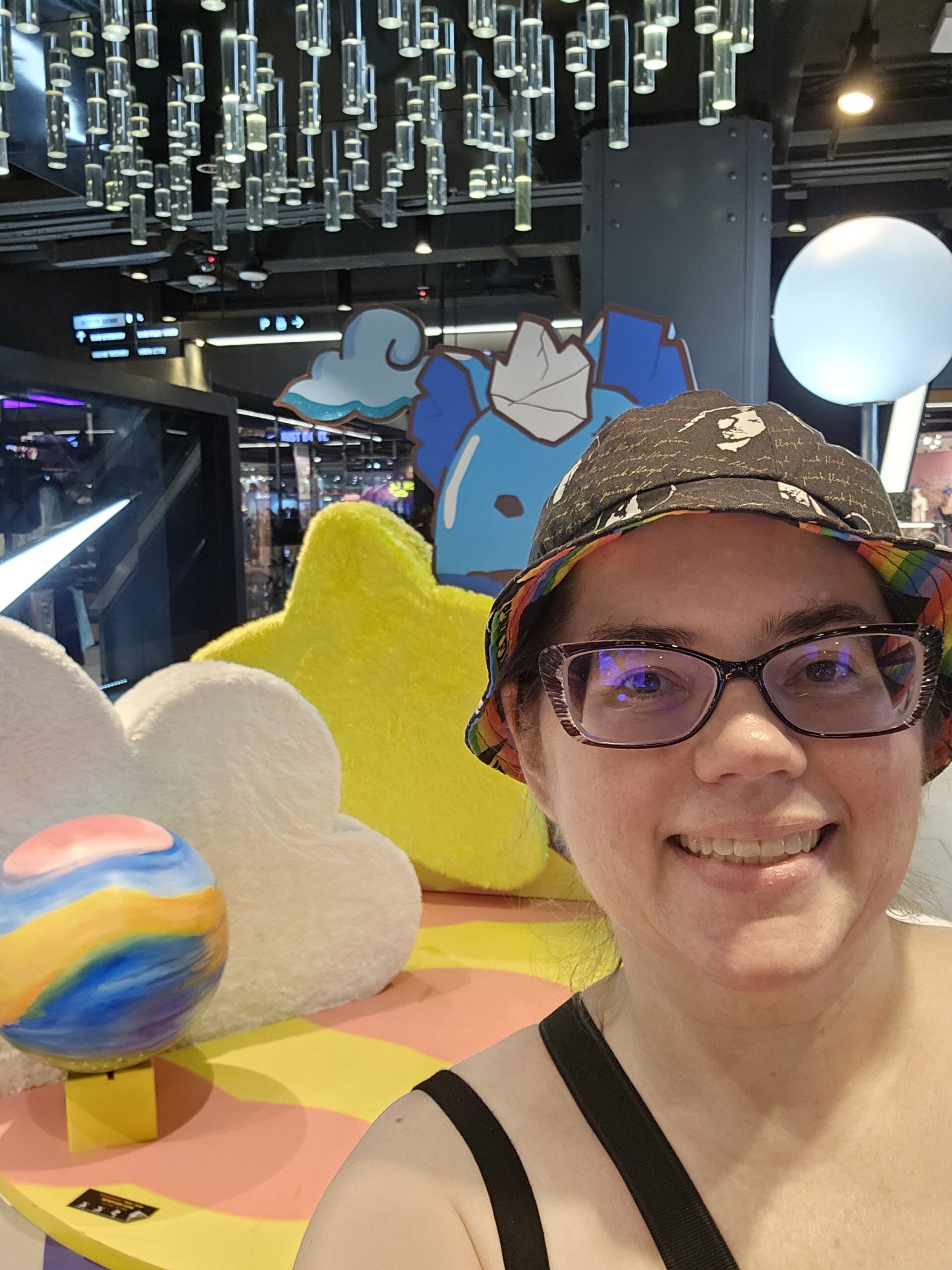 Stephanie, a white woman with glasses and wearing a black bucket hat, standing and smiling in front of some colorful furniture inside a mall.