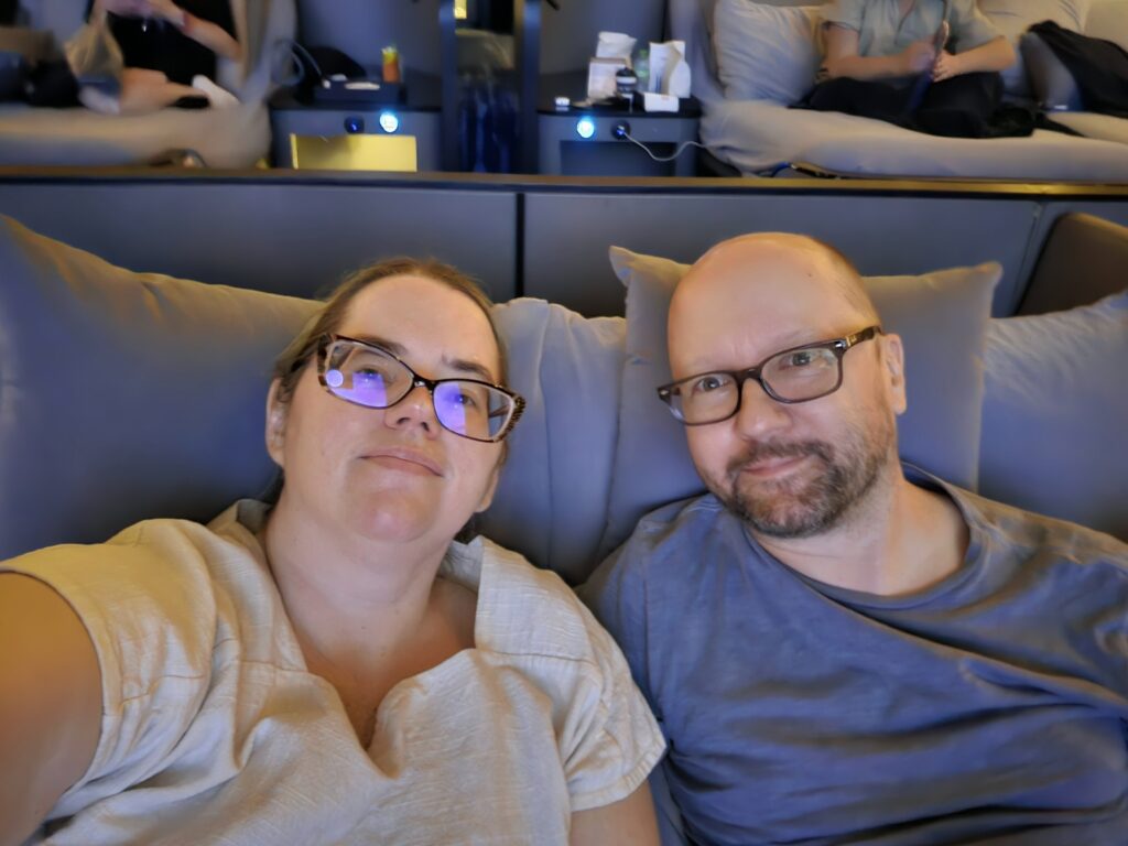 Stephanie and John, a white middle-aged couple wearing glasses. Stephanie has blue hair and is wearing a white linen shirt. John is bald with a beard and is wearing a blue shirt. They are in a lounging bed in a movie theater.