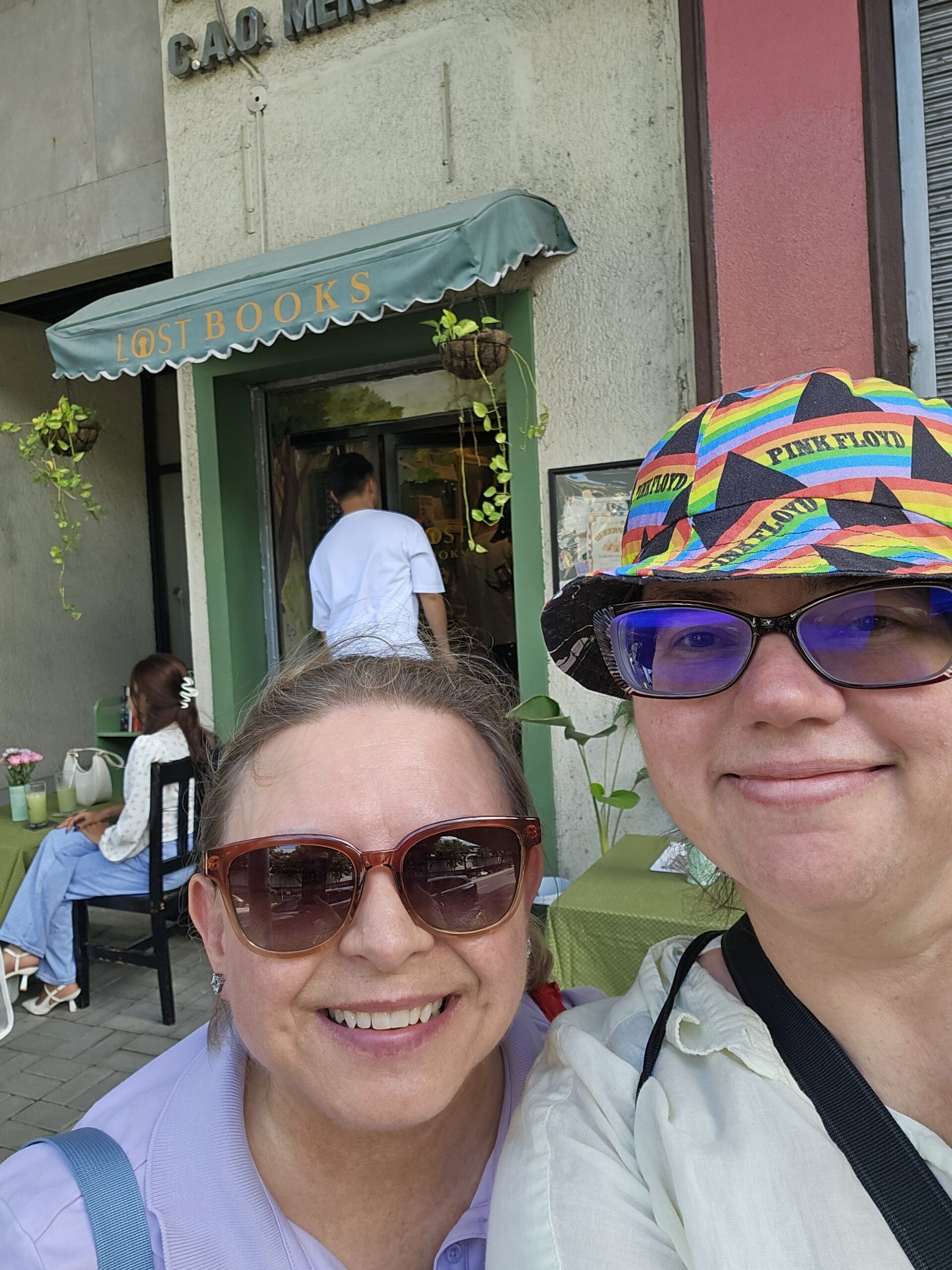 Suzzane (left) and Stephanie (right), two middle aged white women wearing sunglasses, stand in front of Lost Books, a teeny tiny bookshop in Cebu, Philippines.