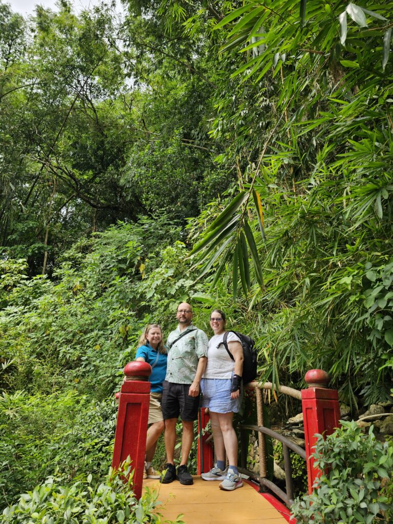 From left to right: Suzanne, John, and Stephanie, three middle-aged white people, standing on a red bridge surrounded by greenery.