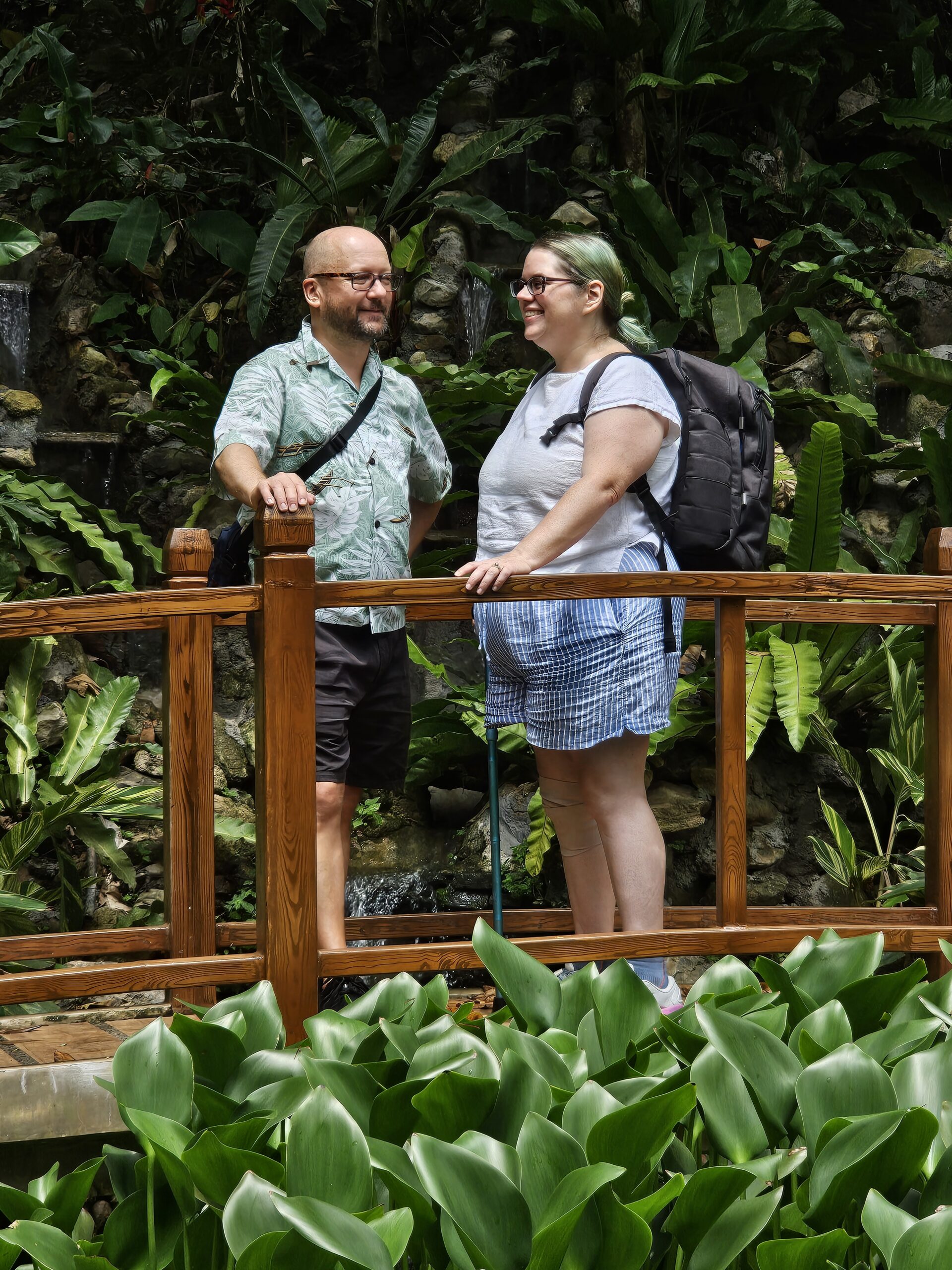 John (left) and Stephanie (right), a middle aged couple, stand on a bridge in the middle of a garden, looking at each other. This pose is reminiscent of their wedding photos.