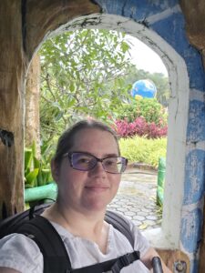 Stephanie, a middle aged white woman wearing glasses, stands in front of an arched window. Behind her is a beautiful garden in the Philippines.