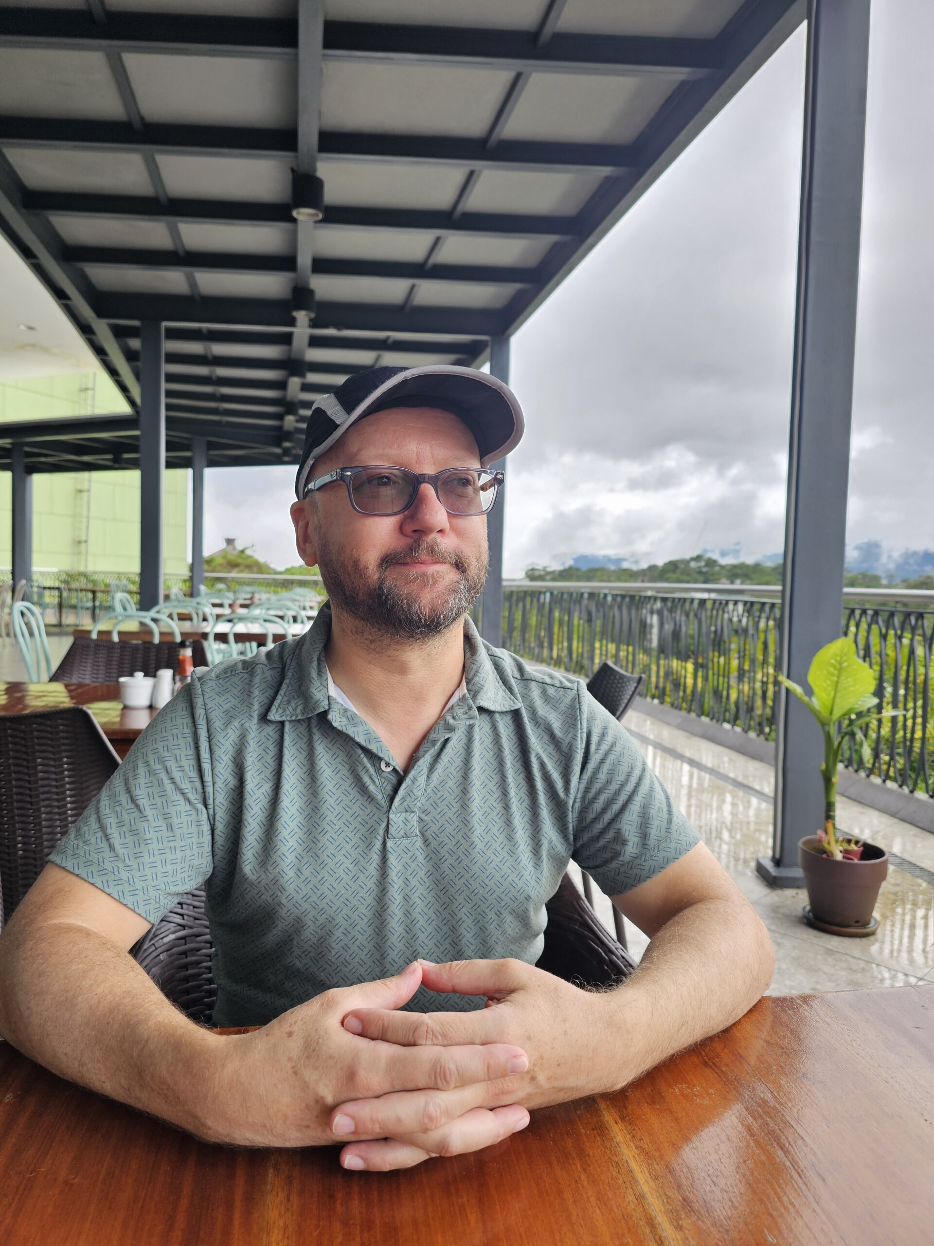 John, a middle aged white man with a beard wearing sunglasses, a billed hat, and a green collared shirt, sits at a table in a Bagiao restaurant outdoors. Storm clouds behind him predict what is about to happen in the next 5-10 minutes.