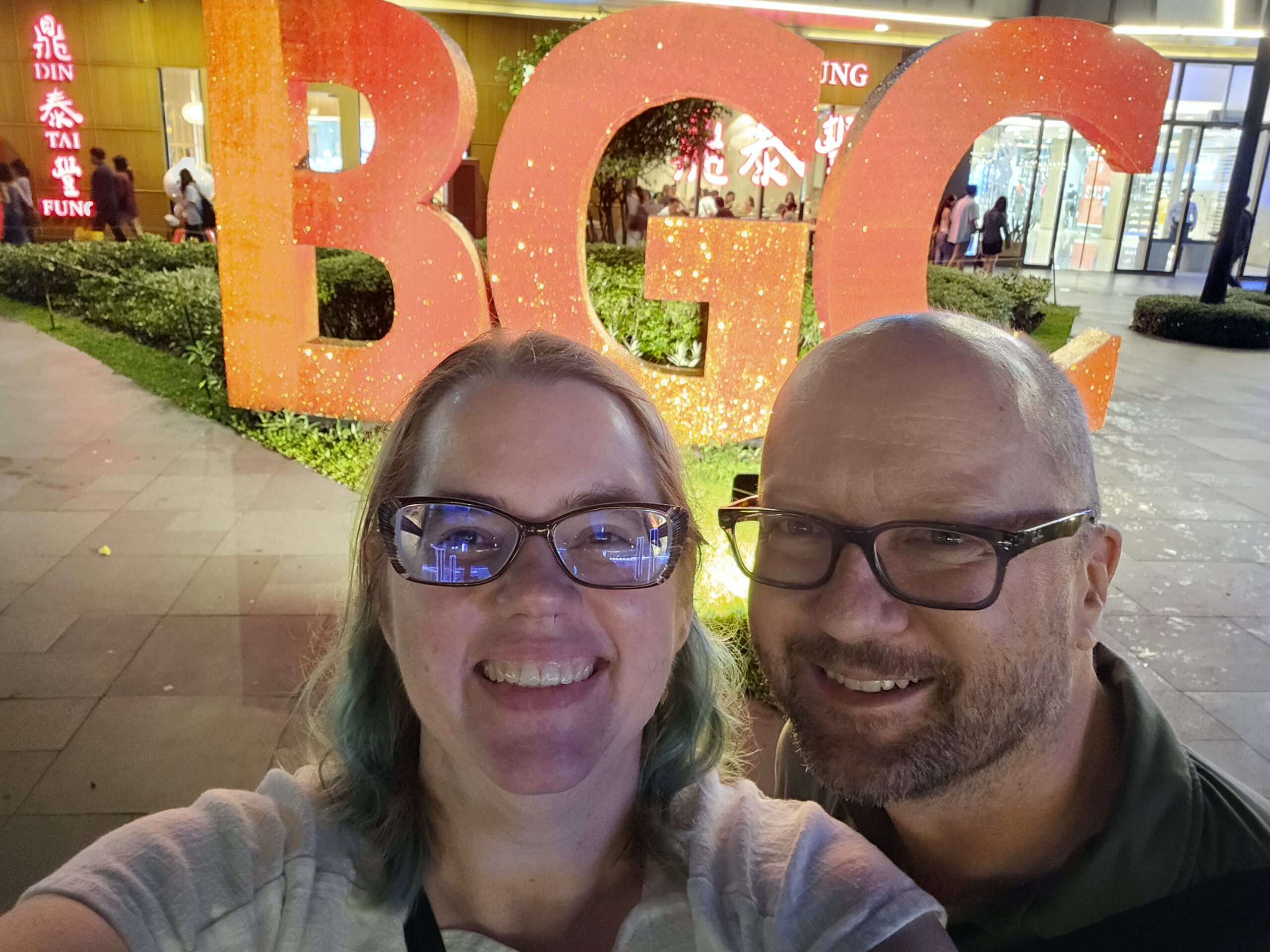 Stephanie (left) and John (right), a middle aged white couple wearing glasses, pose in front of a large orange sign that says "BGC."