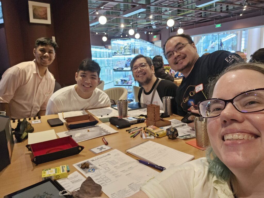 A group of tabletop gamers (mostly male Filipinos, plus Stephanie, a middle-aged white woman) playing Mythic Bastionland at a game store near Manila