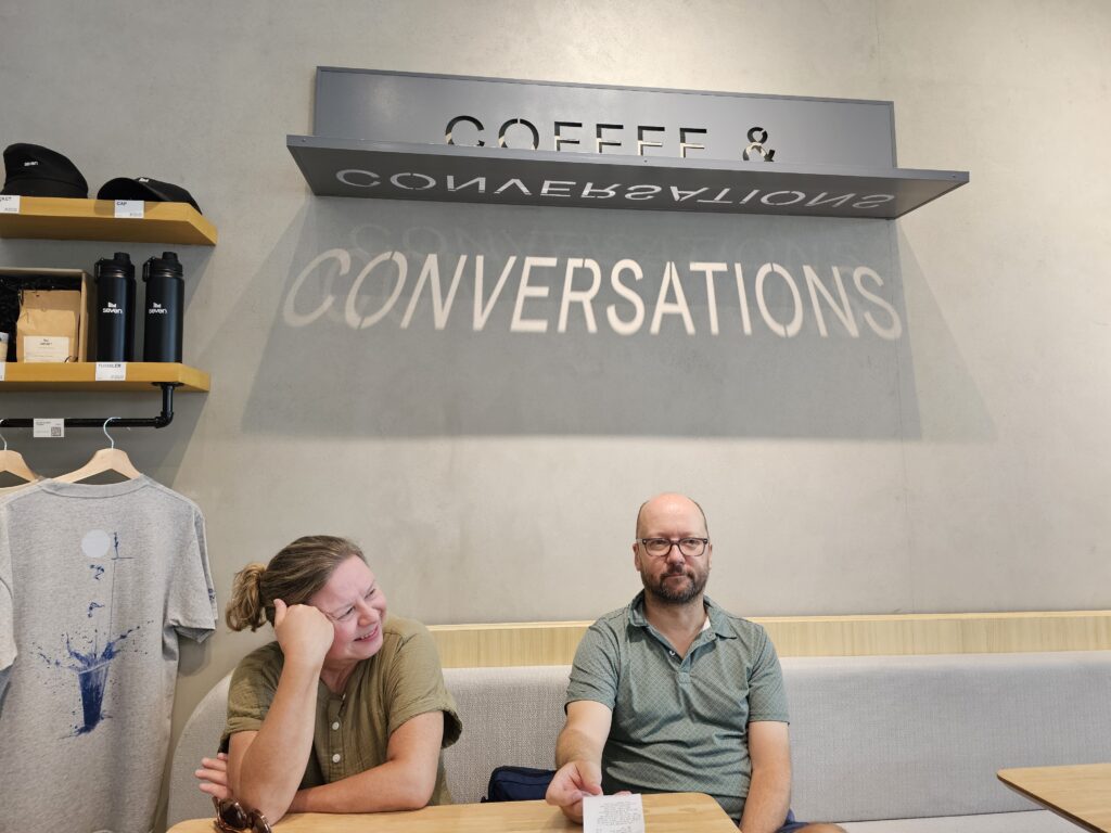 Suzanne (right) and John (left), two middle aged white people, sit underneath a sign that reads "Coffee &" beneath the sign, as a trick of shadow and light, the word "Conversations" is projected onto the wall.