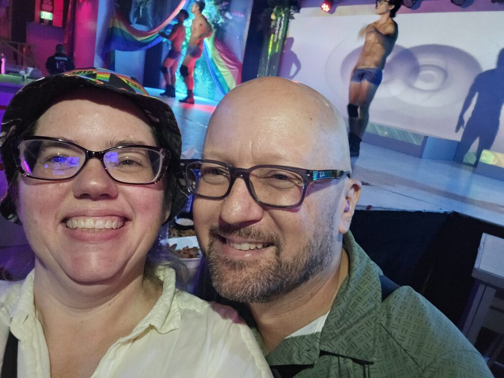 Stephanie and John, a white middle-aged couple in glasses. She is wearing a hat and white collared shirt. He is wearing a green collared shirt. Behind them, a stage has numerous male dancer wearing few clothing, as they are at a queer/drag show.