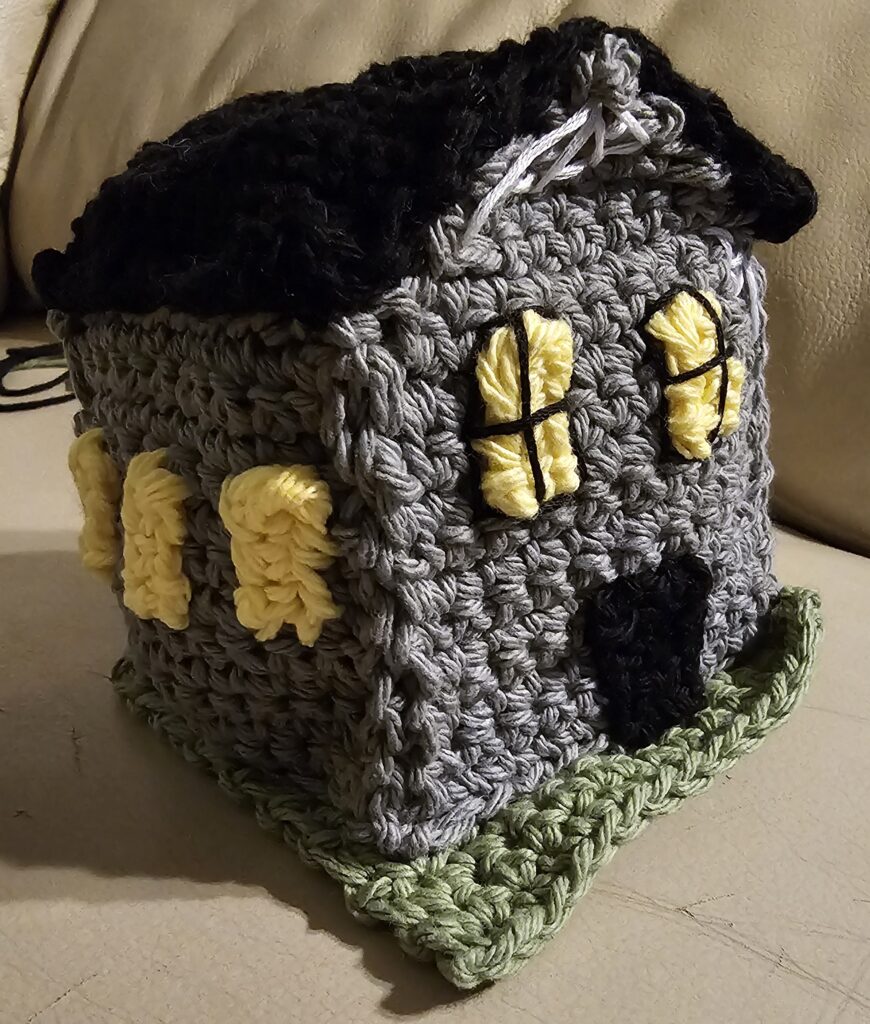 A small grey crocheted house with yellow windows, black roof and front door, and white spiderwebs in the eaves. It is situated on a dark green crocheted lawn.